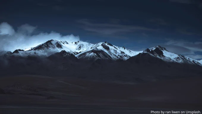 Snow-capped mountain peaks illuminated under a dark, cloudy night sky with a misty valley below.