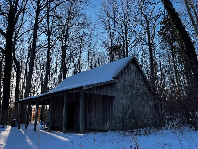 Snow-covered wooden shed against a forest of bare trees, with the sun rising in the background.