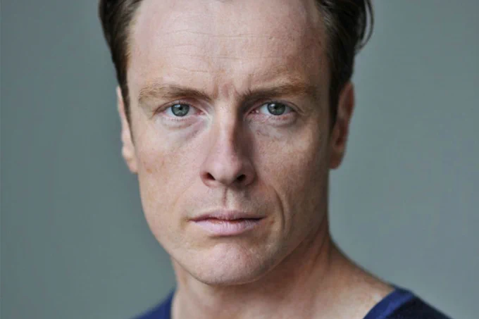 A photo showing a man adopting a neutral expression in front of a grey background 