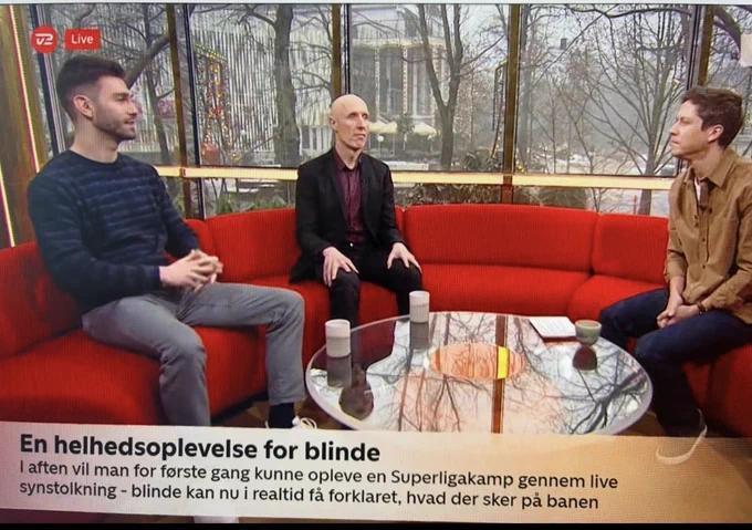 Christian sidder i midten af en rød sofa med synstolker Shelver til venstre og studieværten til højre. På billedet er der en tekstbjælke, hvor der står “En helhedsoplevelse for blinde. I aften vil man for første gang kunne opleve en Superligakamp gennem Love synstolkning - blinde kan nu i realtid få forklaret, hvad der sker på banen”.