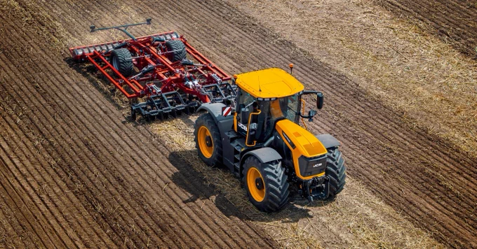 Overhead shot of a yellow JCB tractor pulling a red harrow across a harvested field. The harrow is behind the tractor with its tines in the soil, leaving tracks. The field is mostly brown, with scattered straw.