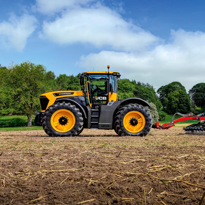 A yellow and black JCB FASTRAC tractor stands on a tilled field under a blue, cloudy sky. Text on the tractor reads \"FASTRAC\" and \"JCB.\" A red implement is attached to the back. Green trees are in the background.