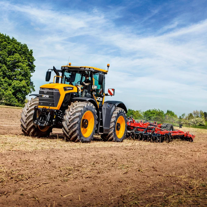 A large yellow and black JCB tractor tills a dirt field on a sunny day with blue skies and fluffy white clouds. The tractor is pulling a red tiller, with a green tree visible on the left side of the image.