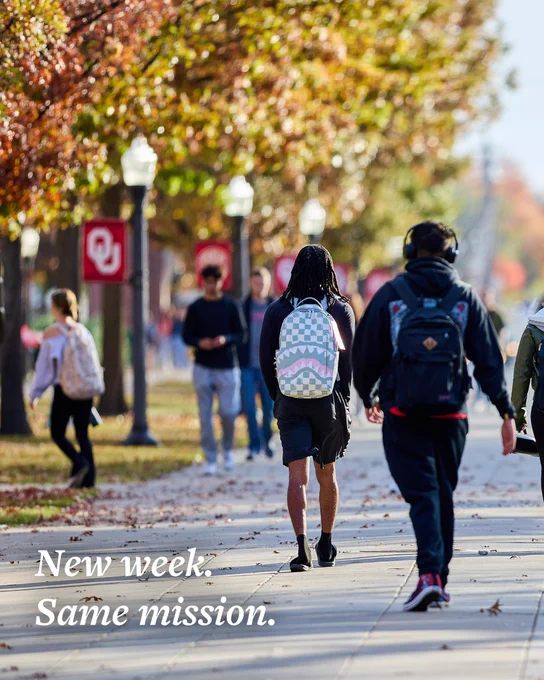 Students with backpacks walk along a tree-lined path on a college campus, with autumn leaves on the ground. A red banner displays the University of Oklahoma logo. Text reads, "New week. Same mission."
