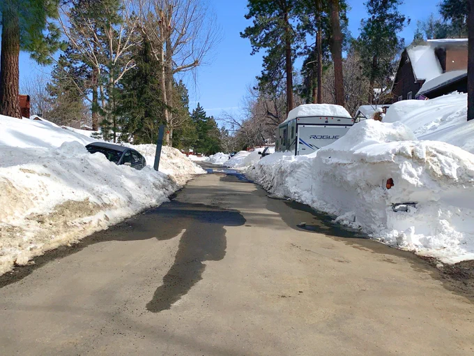 A snow-covered neighborhood street with high snowbanks on either side, some melting on the road. Trees and a Rockwood RV are visible. It is a clear day with a blue sky.