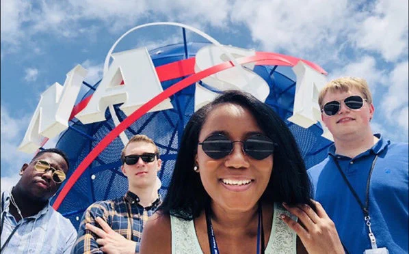 An image of four NASA interns standing in front of a three-dimensional NASA meatball insignia outdoors. The background of the scene is a blue sky filled with diffuse white clouds. Credit: NASA