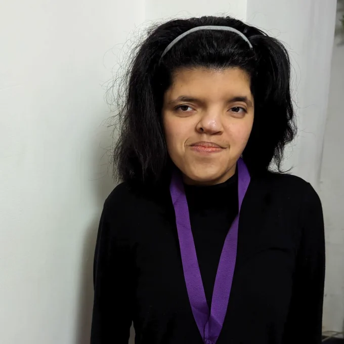 Sneha wears a black long-sleeved dress and has a medal on. Her thick, dark, shoulder-length hair is pushed back by a hair/ head band.