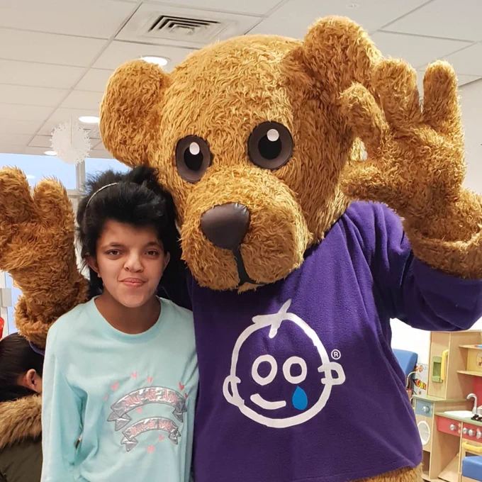 Sneha at Great Ormond Street hospital, next to their mascot, Barnaby bear. She's younger in this picture, with cropped hair.