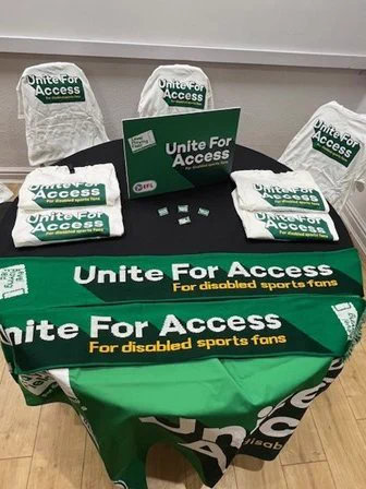A table with chairs and a green tablecloth with badges and a green 'Unite for Access' sign.