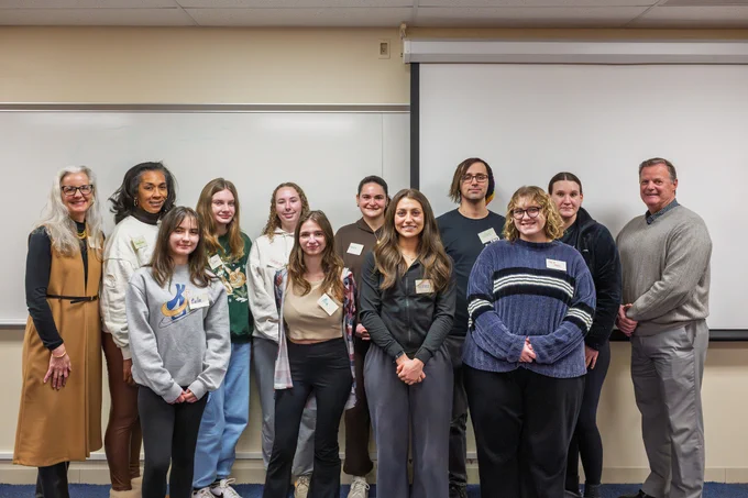 A group of eleven people standing together in a classroom in front of a whiteboard, smiling at the camera. They appear to be casually dressed and wear name tags. The room has beige walls and blue carpeting.