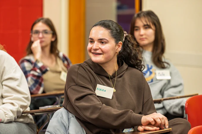 A candid image of a young college student in a brown hoodie smiling and wearing a name tag.