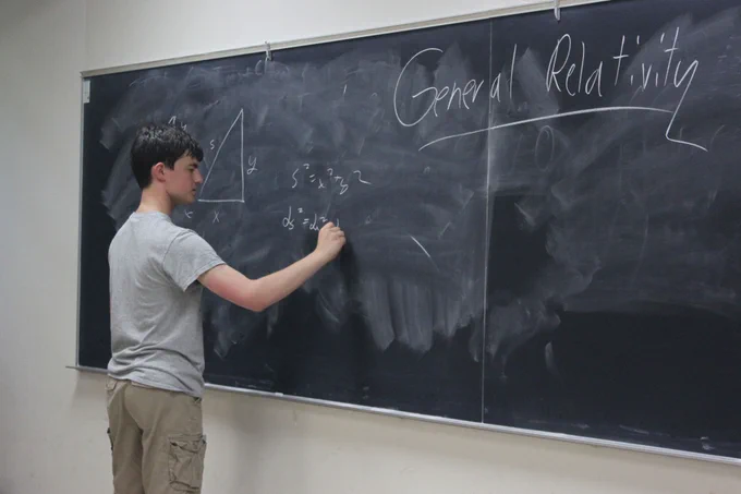 A young Nathan Wagner in high school solving a math equation on a chalkboard. He's wearing a gray t-shirt and khaki cargo shorts.