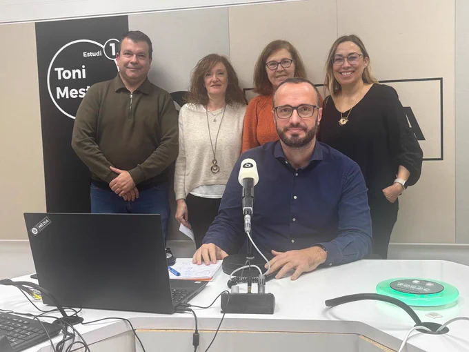 Fotografía en el estudio de radio "Toni Mestre" de À Punt. En primer plano, un hombre sentado frente a un micrófono y un ordenador portátil. Detrás de él, de pie, cuatro personas (un hombre y tres mujeres) sonríen a cámara en un ambiente distendido y profesional. El fondo muestra el logotipo del estudio de radio en la pared