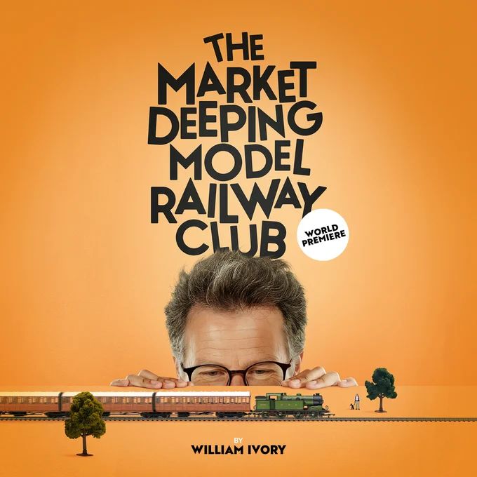A table with a model train running across it, with two model trees, and a model man with a dog. A man with grey hair and glasses is peeking over the edge of the table with smiley eyes. 