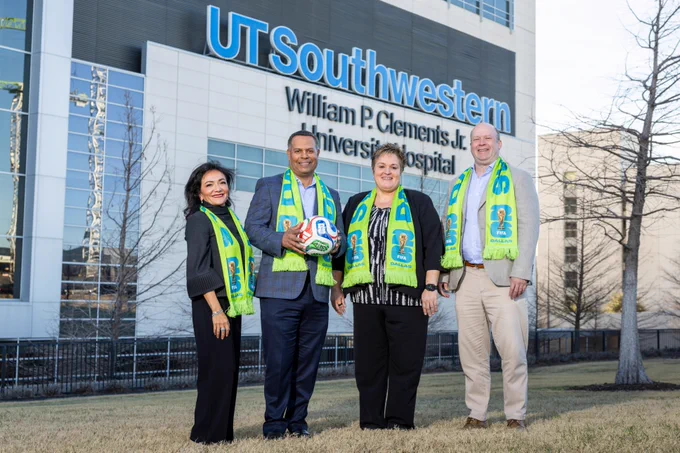 Four people stand outside UT Southwestern William P. Clements Jr. University Hospital, each wearing a green and blue event scarf. One person holds a soccer ball.