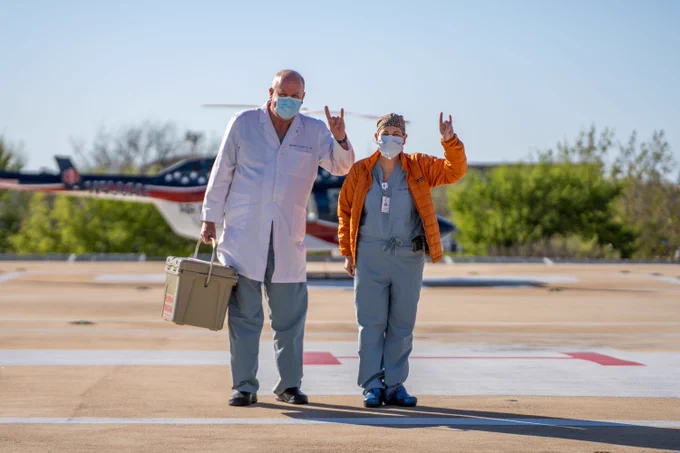 Two medical professionals wearing masks stand on a helipad. One holds a cooler. They both display the Hook 'Em Horns hand sign. A helicopter is visible in the background.