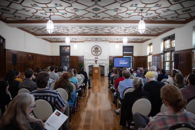 A person speaks at a podium in Beth Moor Lounge, as audience members look on.