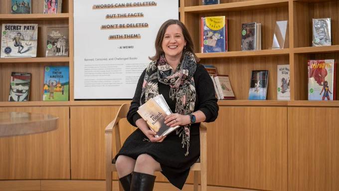 A person sits smiling on a wooden chair, holding a stack of books. There is a large poster in the background on a wall on wooden shelves. 