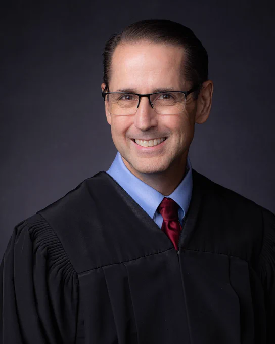 Judge David Zulfa, wearing glasses and a judicial robe, smiling against a dark background.