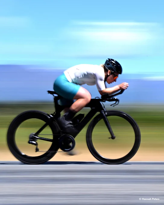 A photo of a woman wearing a white and blue cycling outfit, sunglasses and a helmet riding a black bike on a paved road, leaning forward with motion blur in the background. In the lower right-hand corner, text reads \u201C\u00A9 Hannah Peters.\u201D 