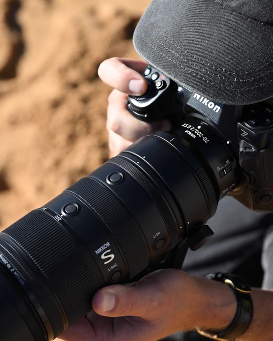 A photo of a pair of hands holding a Nikon Z series mirrorless camera with a NIKKOR Z 70\u2013200mm f/2.8 VR S II lens attached. The photographer, wearing a gray cap, looks through the viewfinder of the camera and has one finger on the shutter button. The golden sand in the background is softly blurred. 