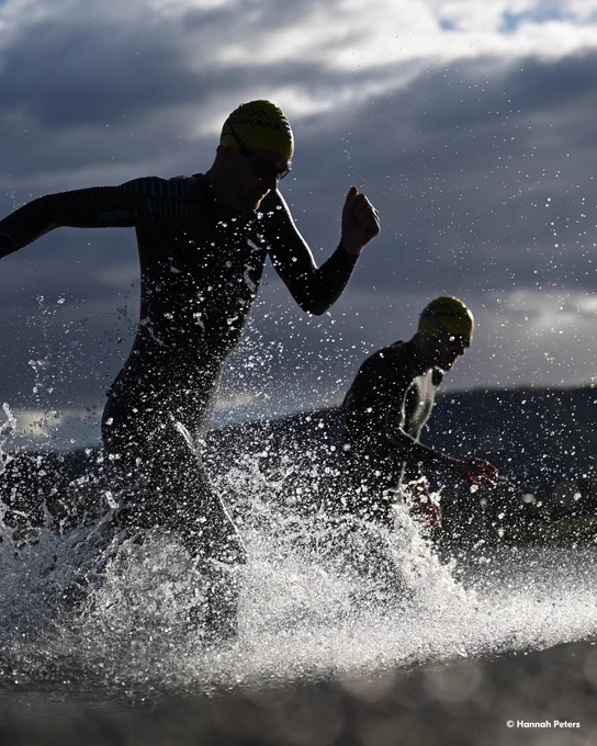 A photo of two people moving through shallow ocean water, splashing as they run under dark, cloudy skies. They are both wearing black wetsuits, swim caps, and goggles. In the lower right-hand corner, text reads \u201C\u00A9 Hannah Peters.\u201D 