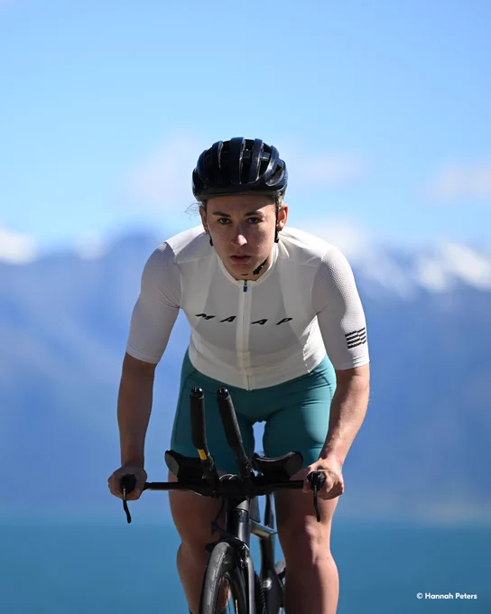 A photo of a woman riding a road bike toward the camera against a bright blue sky and distant mountains, her face in sharp focus. She is wearing a black helmet with a white and blue cycling outfit. In the lower right-hand corner, text reads \u201C\u00A9 Hannah Peters.\u201D 
