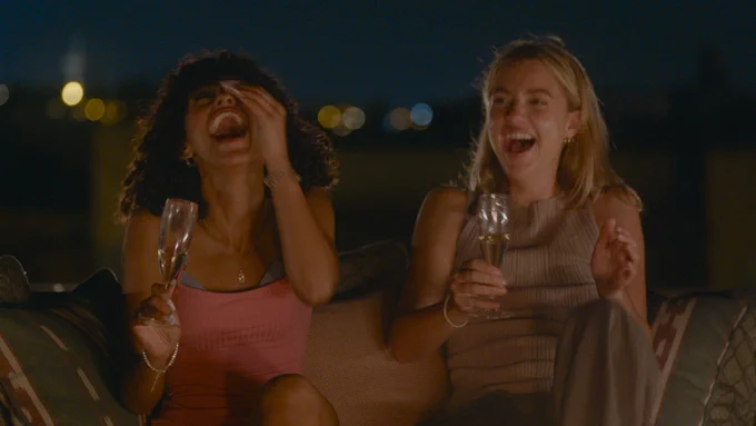 Two women sit outdoors at night laughing and holding champagne glasses, city lights glowing softly behind them.