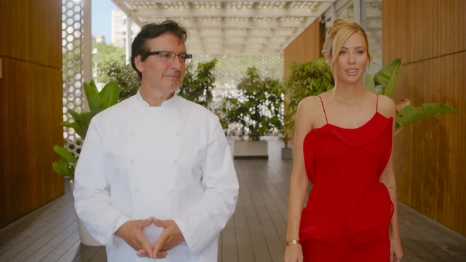 A chef (Jean-Christophe Novelli) and the woman in the red dress (Olivia Attwood) walk side by side through a modern outdoor corridor lined with plants and wooden paneling.