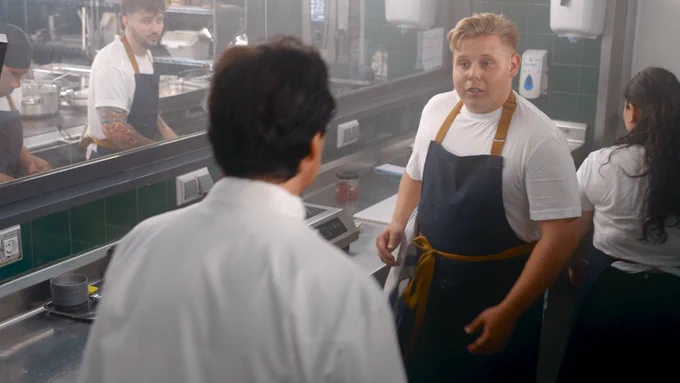 In a busy restaurant kitchen, a young chef in an apron speaks to Jean-Christophe Novelli while other staff work in the background.