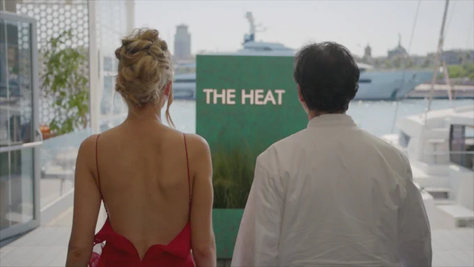 A woman in a backless red dress (Olivia Attwood) and a chef in a white (Jean-Christophe Novelli) jacket stand on a waterfront terrace facing a green sign that reads \u201CTHE HEAT,\u201D with yachts and a city skyline in the background.
