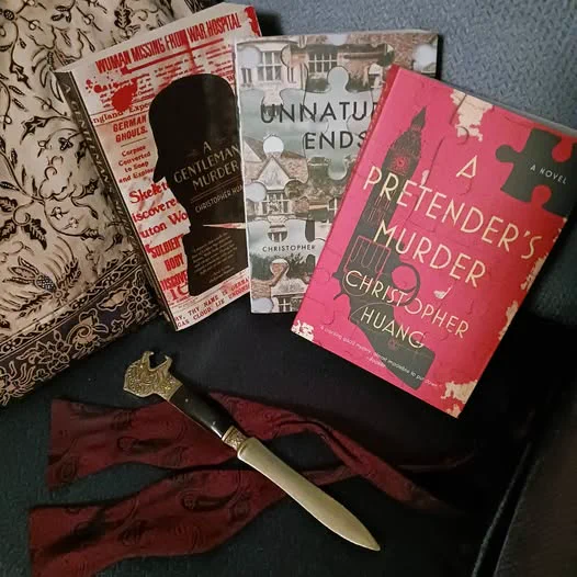 Three books nestled in a blue armchair with a sepia-patterned cushion: A Gentleman's Murder, Unnatural Ends, and A Pretender's Murder. Before them, a dagger-like letter opener with a lion's head handle lies on the coils of an undone burgundy bowtie.