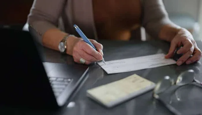 Person writing a check with a pen on a table next to a laptop, checkbook, and glasses.