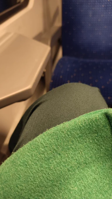 mein Knie in dunkelgrüner Hose, darüber etwas heller der grüne Mantel. unscharf im Hintergrund ein Zug Sitzplatz und das Minitischli in einer SBahn