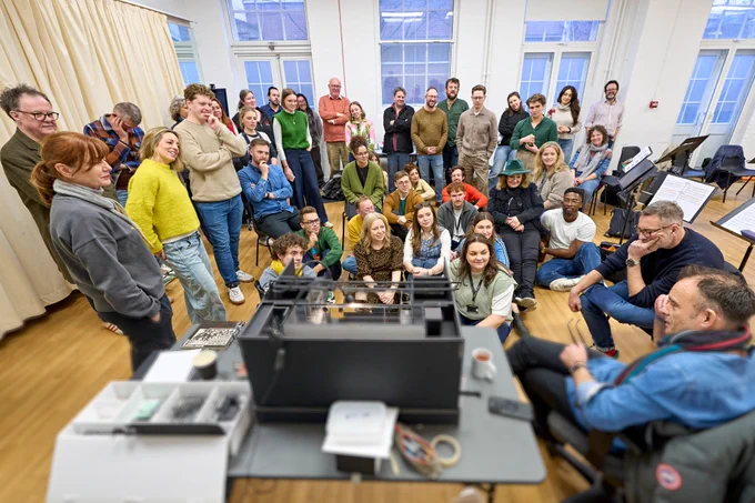A photo showing a group of performers huddle into a rehearsal oom to look at a model box together 