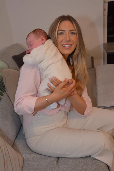 A woman seated on a sofa, holding a newborn baby upright against her shoulder.