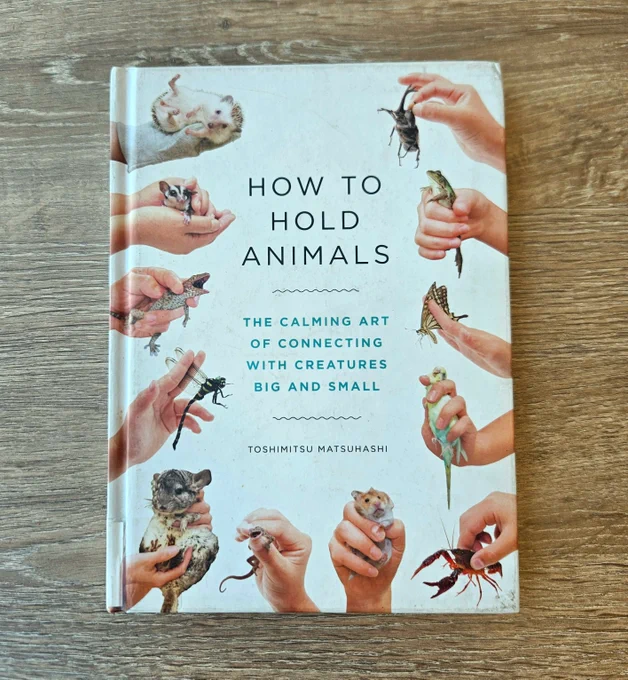 A book cover features various hands gently holding different animals, highlighting techniques for connecting with creatures of all sizes. How to Hold Animals - The calming art of connecting with creatures great and small. By Toshimitsu Matsuhashi. 