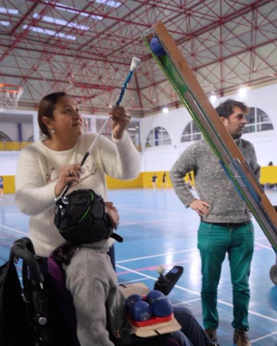 Persona usuaria en silla de ruedas utilizando un puntero de licornio para accionar una rampa de boccia con la asistencia de una mujer. Otra persona permanece de pie observando en un polideportivo.