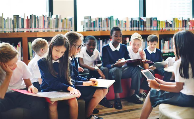A group of students are gathered in a library, sitting on cushions and reading books. The background features shelves filled with colourful books.
