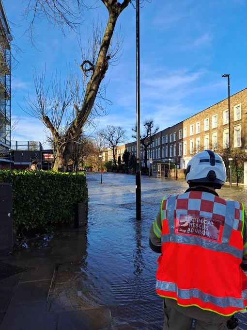 A firefighter in a hi vis vest on the right of the image foreground. In the background, flooding in front of a row of terraced houses.