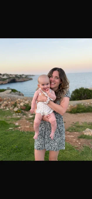 Sally standing outdoors near a coastline, holding a her daughter, Nellie. Sally is wearing a patterned dress and Nellie is dressed in a light coloured outfit.