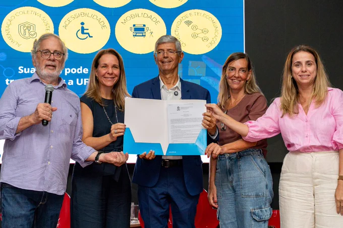 Fotografia durante o lançamento do edital do I Concurso de Soluções Inovadoras da Universidade Federal do Rio de Janeiro (UFRJ). No centro da imagem, o reitor Roberto Medronho segura um documento aberto, simbolizando oficialmente o lançamento. Ao seu lado, também participam do registro, da esquerda para a direita: Fernando Peregrino, pró-reitor de Gestão e Governança, que aparece com um microfone, sugerindo  momento de fala ou apresentação, Lilian Melo Barreto, diretora do Cenpes, Tatiana Sampaio, professora da UFRJ, e Fabiana Valéria da Fonseca, chefe de gabinete da Reitoria. O grupo posa de forma alinhada e sorridente, reforçando o caráter  solene do evento.
