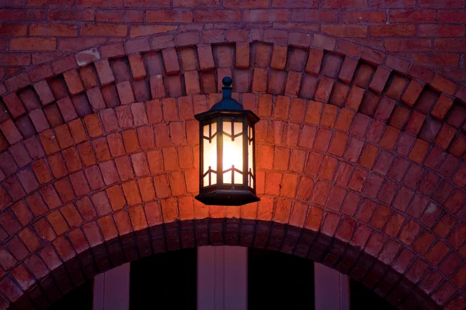 A lantern glows warmly against a brick archway. The bricks are reddish-brown and form a curved structure. The lantern is dark and ornate, casting a soft light.