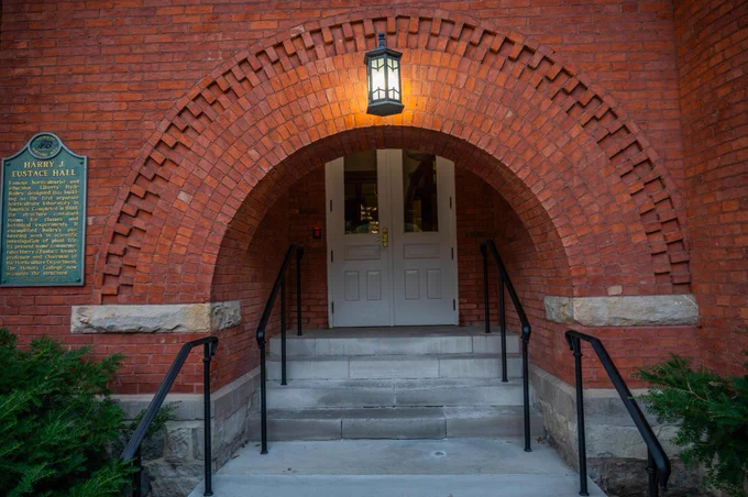A brick arched entrance with steps and black handrails leads to double doors. A lit lantern hangs above. A historical marker reads \"Harry J. Eustace Hall: Famous horticulturist and educator, text@1...The Honors College now occupies the structure.\"