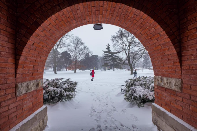 Through a brick arch, a snowy park with bare trees is visible. Two people walk in the distance, leaving footprints in the snow-covered ground. Snow-laden bushes flank the archway.