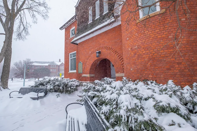 Snowy scene featuring a red brick building with an arched entrance. Benches and bushes in the foreground are covered in snow. A light snowfall blurs the background building.