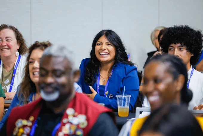 A group of Lions club members sitting in a conference room, smiling and listening. One female Lions member in focus wears a blue blazer, and others are wearing casual attire. The environment appears lively and engaging. She is smiling and learning. 