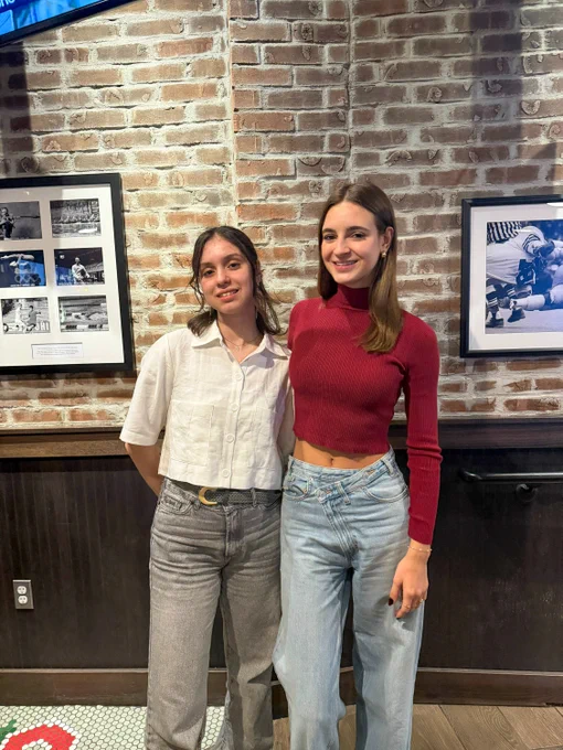 Two individuals are standing in front of a brick wall adorned with framed black and white photographs. The person on the left is wearing a white shirt and jeans, while the person on the right is in a red long-sleeve top and light blue jeans. They are posing casually and smiling.