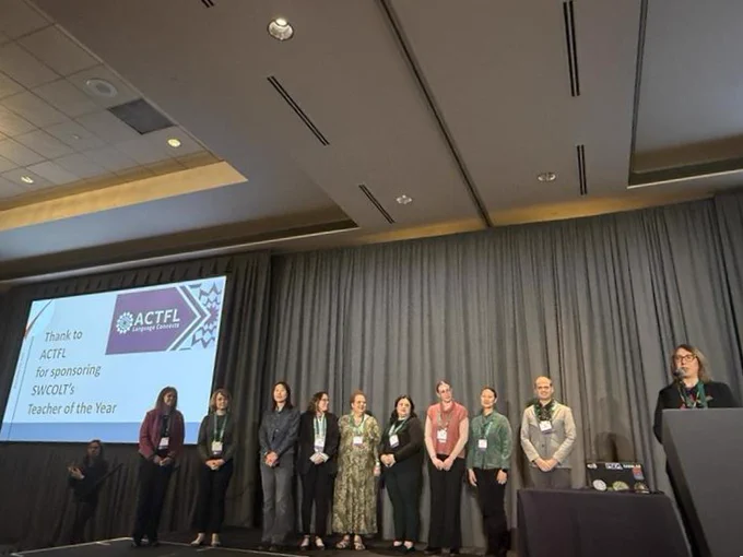 A group of people stand on a stage in front of a curtain. They are in a line, facing an audience. A screen behind them displays the message: "Thanks to ACTFL for sponsoring SWCOLT'S Teacher of the Year." A podium is visible on the right with a speaker addressing the audience.