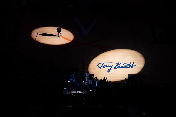 A person stands in a spotlight on a basketball court alongside a larger spotlight that illuminates "Tony Bennett" written on the floor. The setting appears to be a sports arena with minimal ambient lighting.